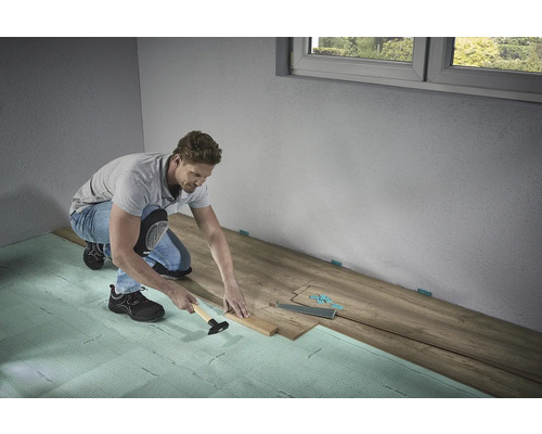 Muž pokládá laminátovou podlahu s kladivem a tloukem na kročejovou izolaci v místnosti.