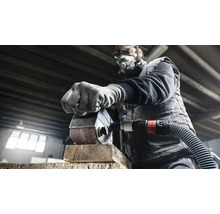 Muž brousí kus dřeva bruskou. Má ochrannou masku a brýle.