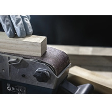 Detailní záběr pásové brusky brousící kus dřeva, obsluhovaný osobou v rukavicích.