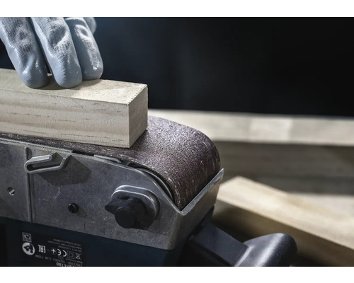 Detailní záběr pásové brusky brousící kus dřeva, obsluhovaný osobou v rukavicích.