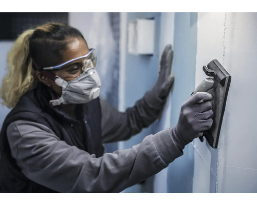 Osoba nosí ochranné pomůcky a hladí stěnu pomocí hladítka.