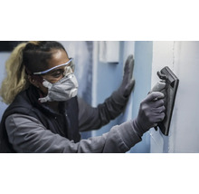 Osoba nosí ochranné pomůcky a používá brusnou desku k broušení stěny.