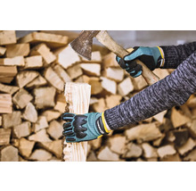 Osoba štípe dříví sekerou a v ochranných rukavicích před hromadou dřeva.