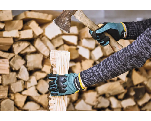 Osoba štípe dříví sekerou a v ochranných rukavicích před hromadou dřeva.