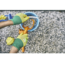 Ruce v zahradních rukavicích plní kyblík lopatkou a kamením, obklopené hračkami na pískoviště.