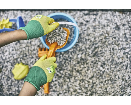 Ruce v zahradních rukavicích plní kyblík lopatkou a kamením, obklopené hračkami na pískoviště.