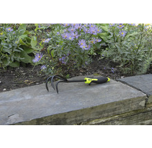 Zahradní kultivátor se třemi hroty a plastovou rukojetí před květinovým záhonem