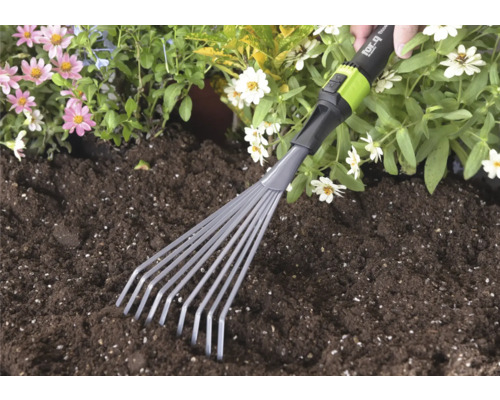 Ruční malé vějířové hrábě for_q používané v tmavé zahradní zemině vedle růžových a bílých květin.