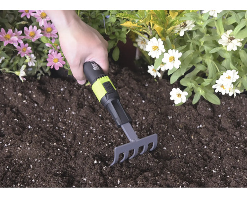 Ruční hrabičky s černo-zelenou rukojetí a for_q Logo používané v tmavé zemině mezi růžovými a bílými květy.