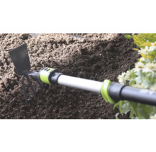 Ruční motyčka s kovovou čepelí a třemi hroty v tmavé zahradní zemině vedle květin.