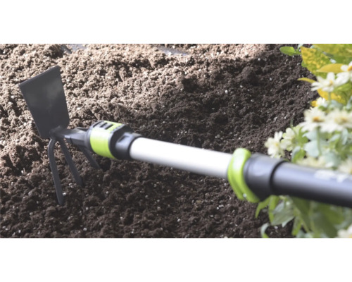 Ruční motyčka s kovovou čepelí a třemi hroty v tmavé zahradní zemině vedle květin.