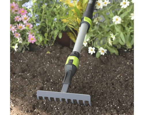 Malé zahradní hrábě s šedou kovovou hlavou a nastavitelnou teleskopickou násadou v zemině před květinami.