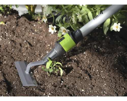 Zahradní plečka s šedou čepelí a zelenou spojkou při odstraňování plevele v tmavé půdě u rostliny.