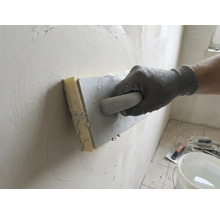 Ruka v pracovní rukavici používá houbové hladítko k vyhlazování omítky na stěně.
