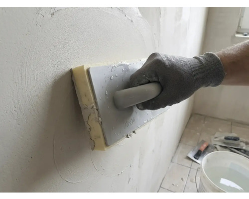Ruka v pracovní rukavici používá houbové hladítko k vyhlazování omítky na stěně.