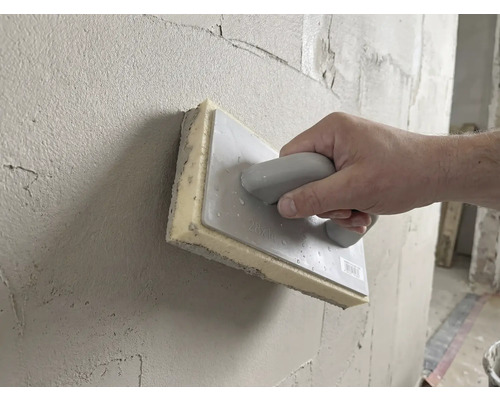 Ruka používá houbové hladítko 28 krát 14 centimetrů k vyhlazování čerstvé omítky na stěně.
