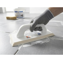 Osoba v ochranné rukavici nanáší pomocí epoxidového hladítka bílou spárovací hmotu na šedou dlažbu.