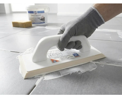 Osoba v ochranné rukavici nanáší pomocí epoxidového hladítka bílou spárovací hmotu na šedou dlažbu.