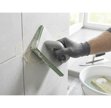 Osoba spáruje nástěnné obklady pomocí spárovacího hladítka. V pozadí je kbelík s vodou, houba a hladítko.