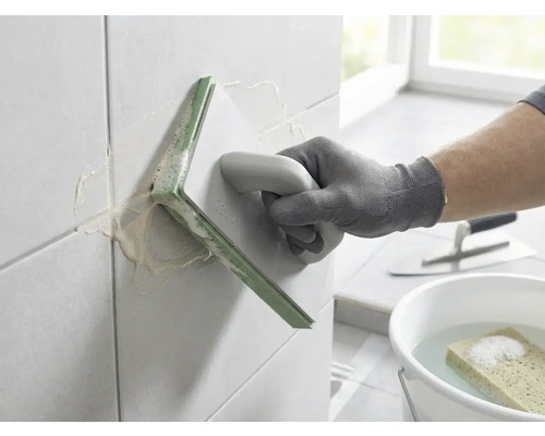 Osoba spáruje nástěnné obklady pomocí spárovacího hladítka. V pozadí je kbelík s vodou, houba a hladítko.