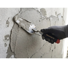 Ruka v pracovní rukavici nanáší maltu na stěnu pomocí zednické lžíce Maurerlob.