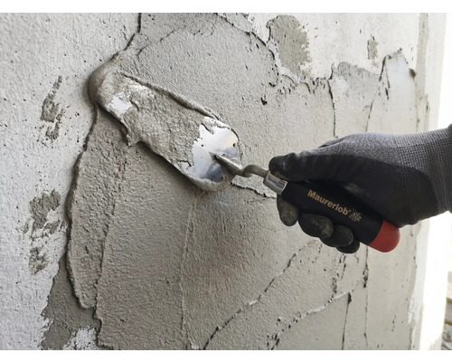 Ruka v pracovní rukavici nanáší maltu na stěnu pomocí zednické lžíce Maurerlob.