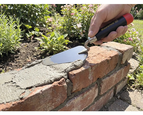 Ruka se zednickou lžící Maurerlob nanáší maltu na cihlovou zídku v zahradě.