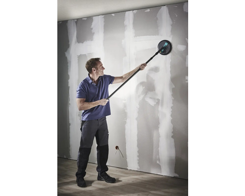 Muž brousí sádrokartonovou stěnu pomocí brusky na sádrokarton s teleskopickou tyčí.