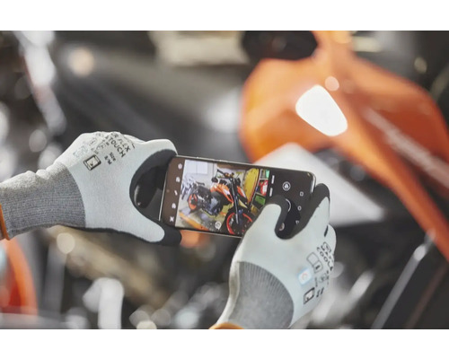 Osoba má na sobě pracovní rukavice s dotykovou funkcí a chytrým telefonem fotografuje motocykl v dílně.
