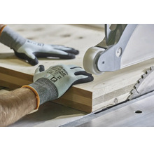 Ruce v pracovních rukavicích vedou dřevěné prkno na stolní kotoučové pile pro přesné opracování dřeva.