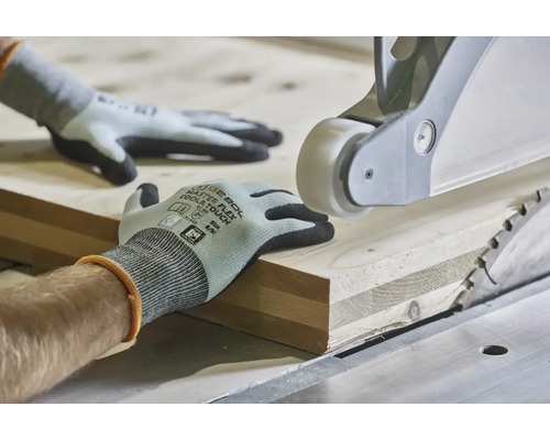 Ruce v pracovních rukavicích vedou dřevěné prkno na stolní kotoučové pile pro přesné opracování dřeva.
