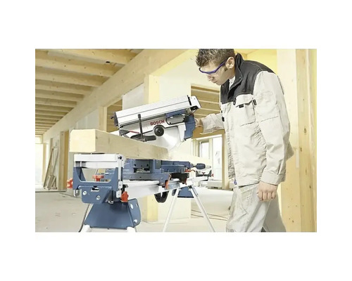 Řemeslník řeže dřevěný trám kapovací a pokosovou pilou Bosch na pracovním stole na světlé stavbě.