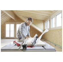 Muž s ochrannými brýlemi řeže dřevěné prkno stolní kotoučovou pilou ve světlé místnosti s dřevěnými trámy a OSB deskami.