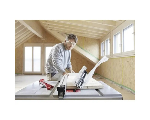 Muž s ochrannými brýlemi řeže dřevěné prkno stolní kotoučovou pilou ve světlé místnosti s dřevěnými trámy a OSB deskami.