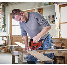 Řemeslník v jasné dílně řeže dřevěné prkno na pracovním stole pomocí akumulátorové přímočaré pily Einhell. Einhell Logo.