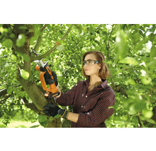 Žena používá akumulátorovou pilu ocasku Worx k prořezávání větví v zahradě. Má ochranné brýle a rukavice.
