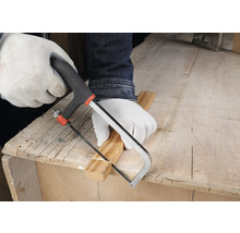 Osoba v bílých ochranných rukavicích řeže dřevěnou lištu malou ruční pilkou na pracovním stole.