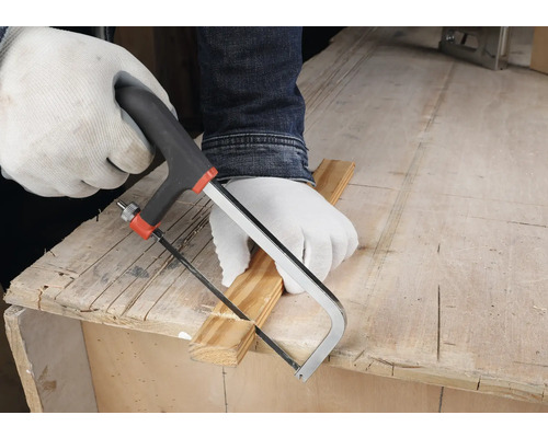 Osoba v bílých ochranných rukavicích řeže dřevěnou lištu malou ruční pilkou na pracovním stole.