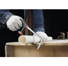 Osoba v rukavicích řeže bílou plastovou trubku malou pilkou na kov na dřevěném pracovním stole.