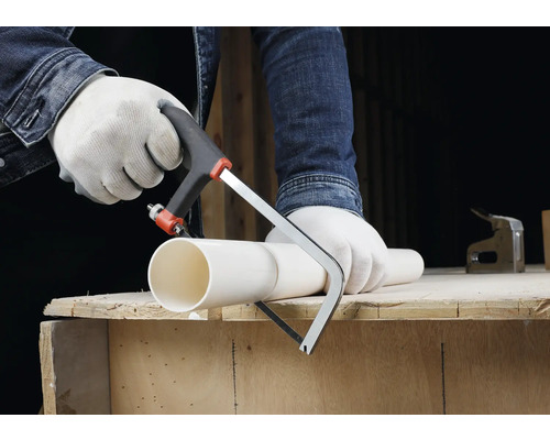 Osoba v rukavicích řeže bílou plastovou trubku malou pilkou na kov na dřevěném pracovním stole.