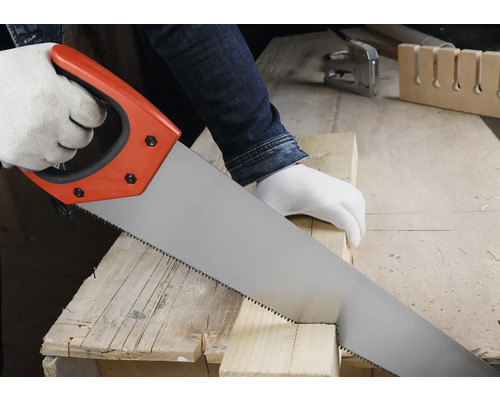 Osoba řeže dřevěný hranol ruční pilou na pracovním stole v jasné dílně.