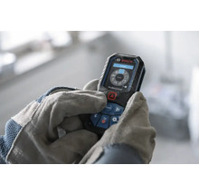 Laserový dálkoměr Bosch Professional ovládaný rukama v pracovních rukavicích, digitální displej s funkcemi měření.