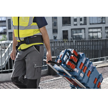 Řemeslník v reflexní vestě táhne transportní vozík naložený kufry na nářadí Bosch L-BOXX a stavebním rádiem na staveništi.