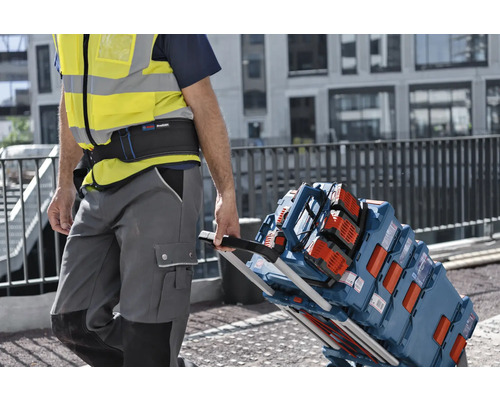 Řemeslník v reflexní vestě táhne transportní vozík naložený kufry na nářadí Bosch L-BOXX a stavebním rádiem na staveništi.