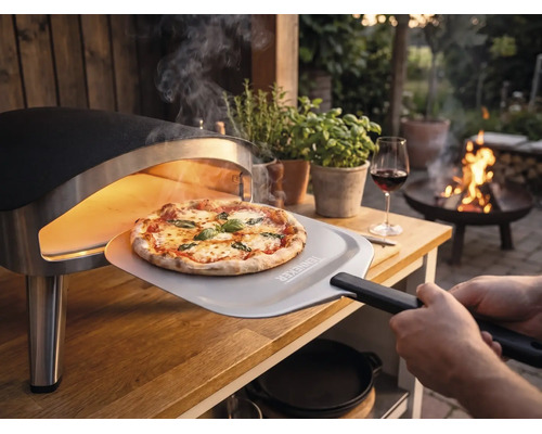 Čerstvě upečená pizza se vyjímá z přenosné pece na pizzu na dřevěném stole na zahradě pomocí lopaty na pizzu s Tenneker Logo.
