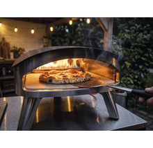 Venkovní nerezová pec na pizzu s hořícími plameny a pizzou na pizzovém kameni, s níž se manipuluje pomocí lopaty na pizzu.