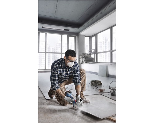 Muž klečí v místnosti a řeže dlaždici bruskou. Má na sobě ochrannou masku a ochranné brýle.