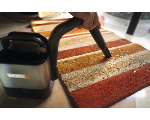 Akumulátorový vysavač se používá na pruhovaném koberci k odstranění malých částic. Logo společnosti Worx.