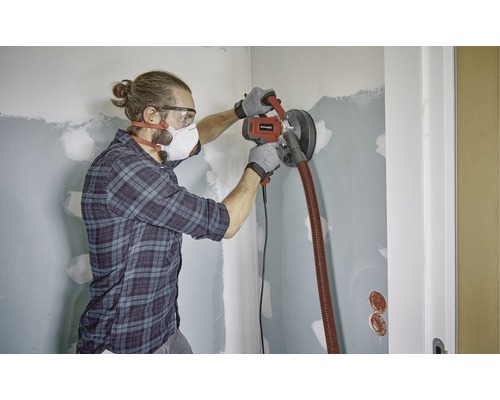 Muž brousí stěnu sádrokartonovou bruskou v místnosti.