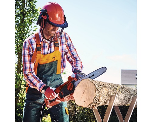 Muž řeže elektrickou řetězovou pilou špalek na kozlíku a má na sobě helmu s obličejovým štítem a chrániče sluchu